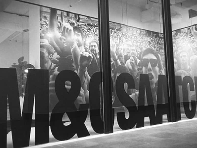 M&C Saatchi branding on office glass door with crowd reflection.