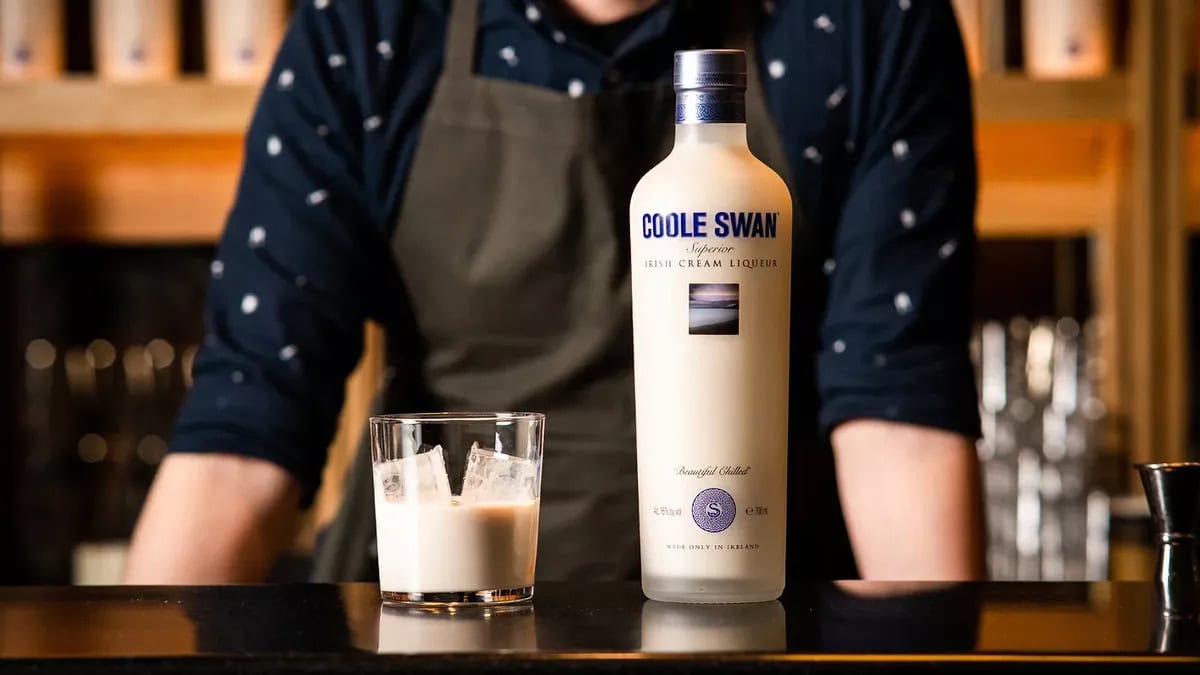 Close-up of Coole Swan bottle and glass on a bar counter, with a bartender in the background.